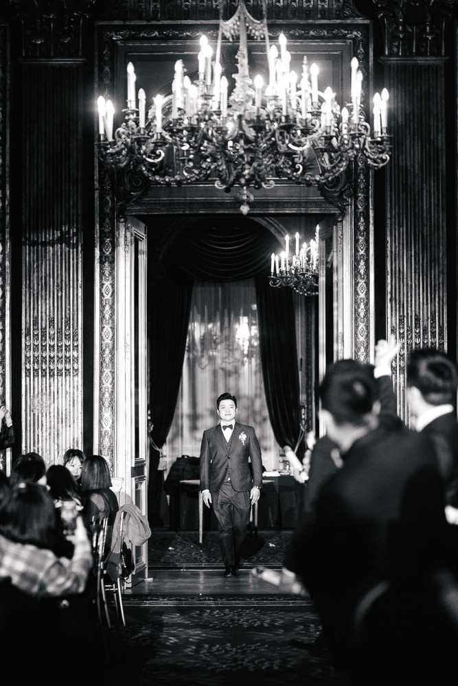This black-and-white image captures the groom making his processional entrance down a central aisle inside a grand, ornate ballroom or palace-style interior. The groom wears a dark three-piece suit with a bow tie and a boutonnière on his lapel, walking toward the camera with a smile. The room features heavily carved dark wood paneling, tall decorative doorframes, draped curtains, and multiple large candelabra-style chandeliers with lit candles that create bright highlights against the dark tones of the image. Seated guests line both sides of the aisle, with approximately 20–30 visible attendees. The composition is a centered wide shot taken from the far end of the aisle, framing the groom symmetrically between the architectural elements and chandelier above. The overall aesthetic is classic and formal, consistent with a historic château or hôtel particulier setting.