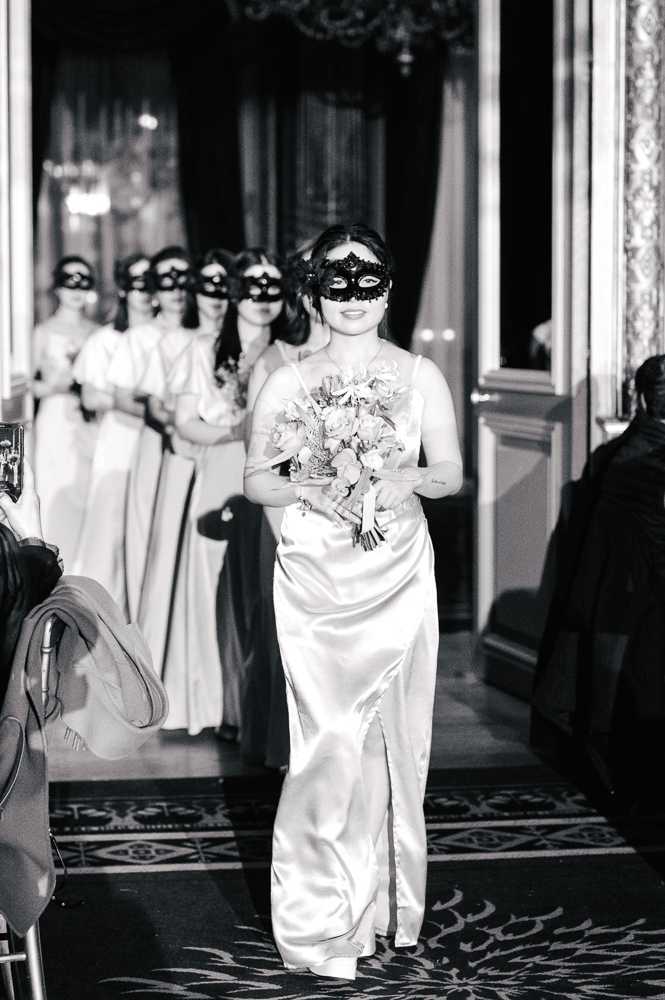 Black-and-white portrait-style shot of a bridal party processional taking place inside an ornate ballroom or grand hall, identifiable by a chandelier, decorative molding, and patterned carpet aisle. The bride leads the procession wearing a satin slip-style gown with spaghetti straps and a front slit, carrying a round bouquet, while approximately six bridesmaids follow behind her in light-colored floor-length gowns. All members of the bridal party, including the bride, are wearing black decorative masquerade-style eye masks, indicating a masquerade-themed wedding. The image is shot at medium distance from a low angle along the aisle, with guests visible on the left edge of the frame, and shows high contrast between the light gowns and the dark masks and background decor.