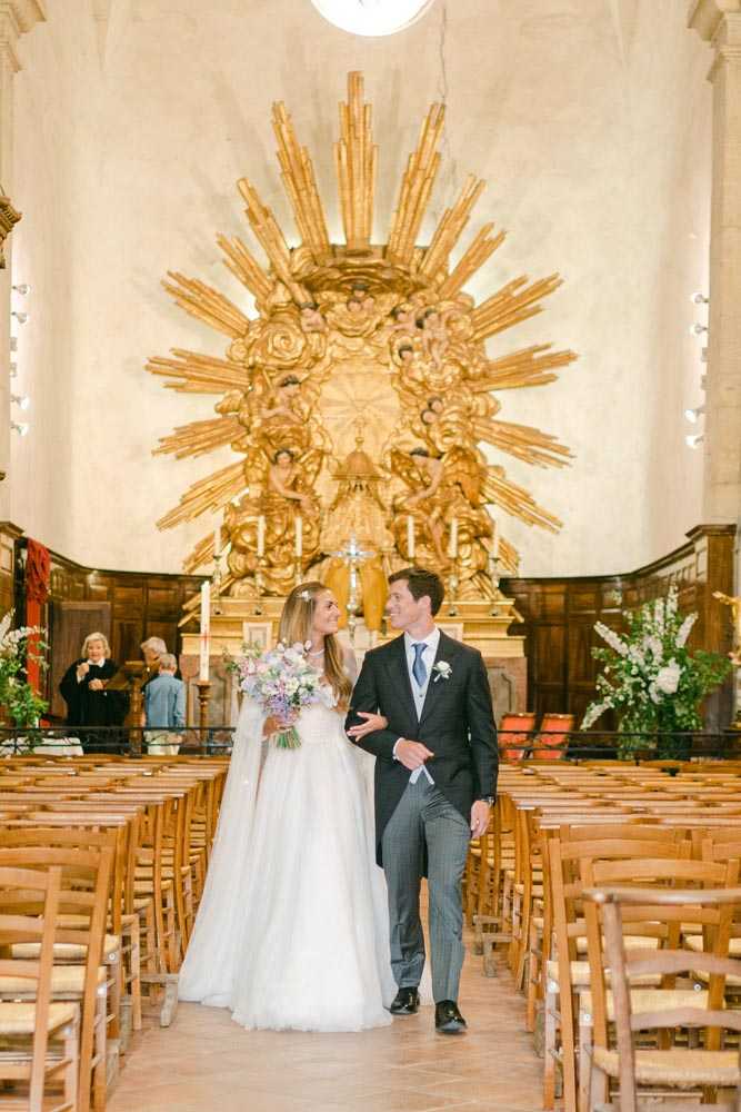 The bride and groom walk back down the aisle together following their church ceremony, photographed in a wide portrait shot. The setting is a classic French stone chapel with an ornate gilded Baroque altarpiece featuring a large sunburst design as the focal backdrop. The bride wears a white tulle ball gown with a long cape veil and carries a loose, garden-style bouquet of lavender, lilac, and soft pink blooms. The groom is dressed in a dark charcoal suit with grey pinstripe trousers, a light blue tie, and a white boutonniere. The couple look at each other smiling as they walk between rows of natural wood rush-seated chairs. Two guests and a celebrant are visible near the altar in the background, along with tall white candles and a large white floral arrangement with greenery flanking the right side of the altar.