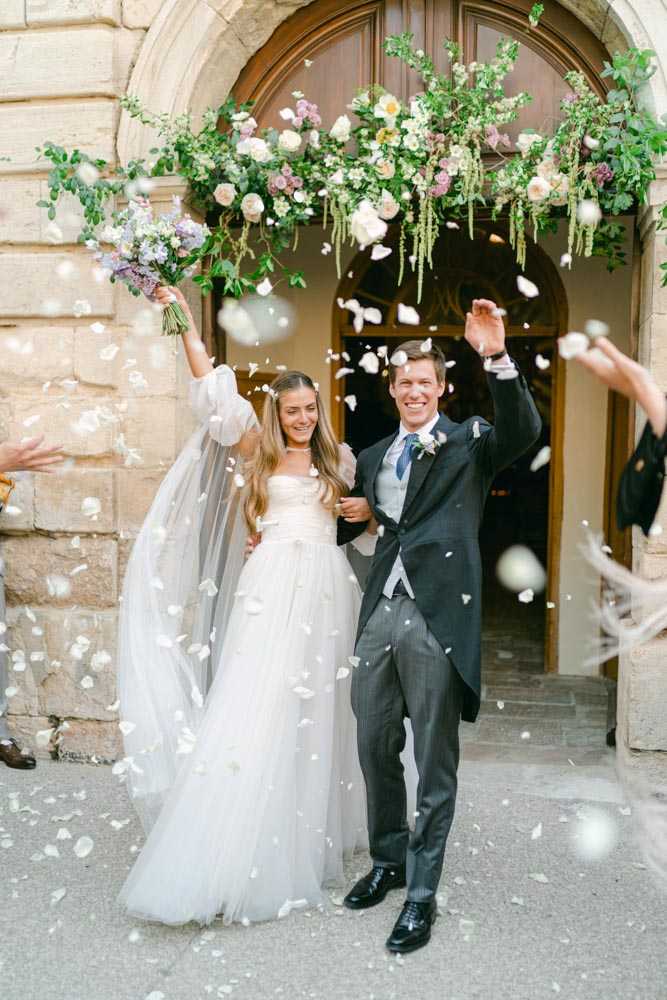The bride and groom are exiting a ceremony venue through an arched stone doorway as guests throw white rose petals around them. The bride wears a white tulle ball gown with puff sleeves and floral appliqué detailing at the bodice, paired with a long veil and a loose hair flower accessory; she holds her bouquet of lavender, lilac, and cream blooms raised in the air. The groom wears a charcoal grey suit with a light grey waistcoat, a navy patterned tie, and a white boutonnière. Above the doorway hangs a large floral installation featuring blush and cream roses, mauve blooms, and trailing greenery. This is a portrait-style mid shot capturing the celebratory moment of the couple's ceremony exit.