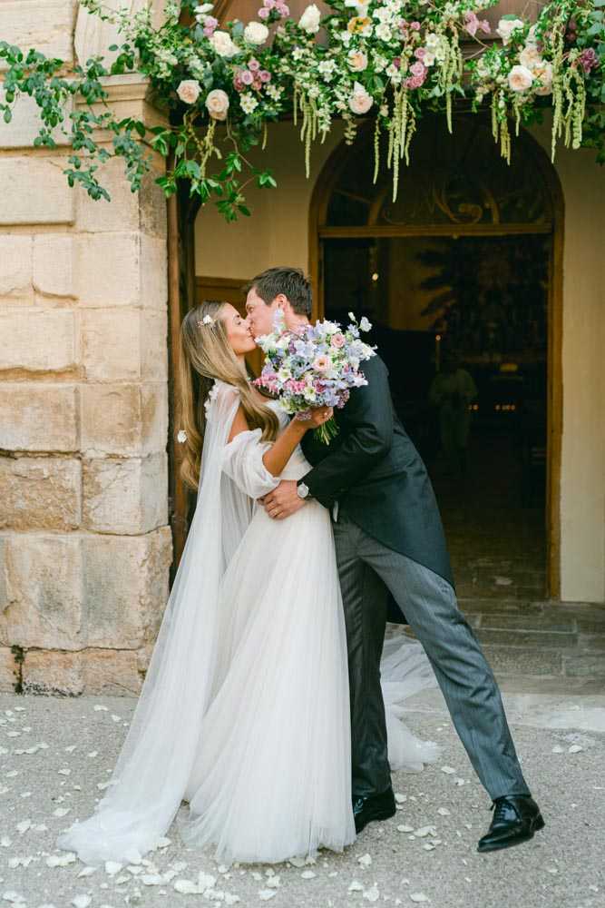 A bride and groom share a kiss just outside the entrance of a stone chateau or manor building, with scattered flower petals on the ground around them. The bride wears a white tulle off-shoulder gown with sheer long sleeves and a flowing layered skirt, her long hair down with small floral pins, and she holds a loose, garden-style bouquet of lavender, blush pink, soft blue, and white blooms including sweet peas and anemones. The groom wears a dark charcoal morning coat with grey striped trousers and black Oxford shoes, and dips the bride slightly as they kiss. Above them hangs a lush floral installation featuring cascading greenery, blush garden roses, mauve flowers, and trailing vines framing the arched doorway, creating a romantic, garden-inspired ceremony exit moment. Portrait shot, taken outdoors in natural light.