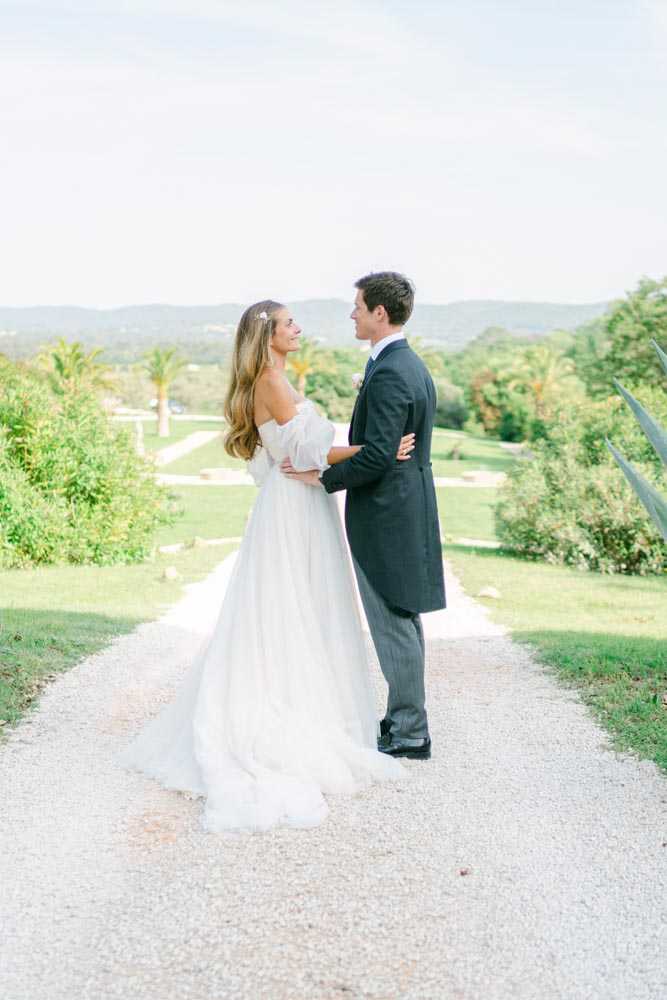 A couple portrait taken outdoors on a gravel driveway lined with manicured lawns, with a tree-covered hillside visible in the background suggesting a southern French property. The bride wears an off-the-shoulder white gown with voluminous puff sleeves and a full skirt with a slight train, her long hair worn down with a small floral hair accessory. The groom wears a charcoal grey morning suit with a white boutonnière. The two face each other in an embrace, the bride smiling up at the groom. The image is a full-length portrait shot with soft, bright natural light and a slightly airy, film-inspired tone.