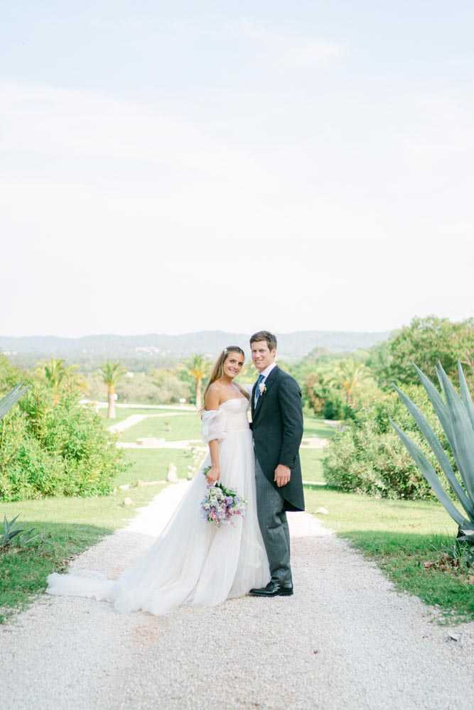 A couple portrait taken outdoors on a gravel pathway with a sweeping garden and tree-lined landscape visible in the background. The bride wears an off-the-shoulder white ballgown with a full tulle skirt and a long train, holding a loosely arranged bouquet of purple, lavender, and pink blooms including what appear to be sweet peas and ranunculus. The groom wears a charcoal grey suit with a light blue tie and a small white floral boutonniere. The two stand close together, both facing the camera, in a classic portrait composition at medium distance. The overall styling is classic and airy, with soft natural lighting.