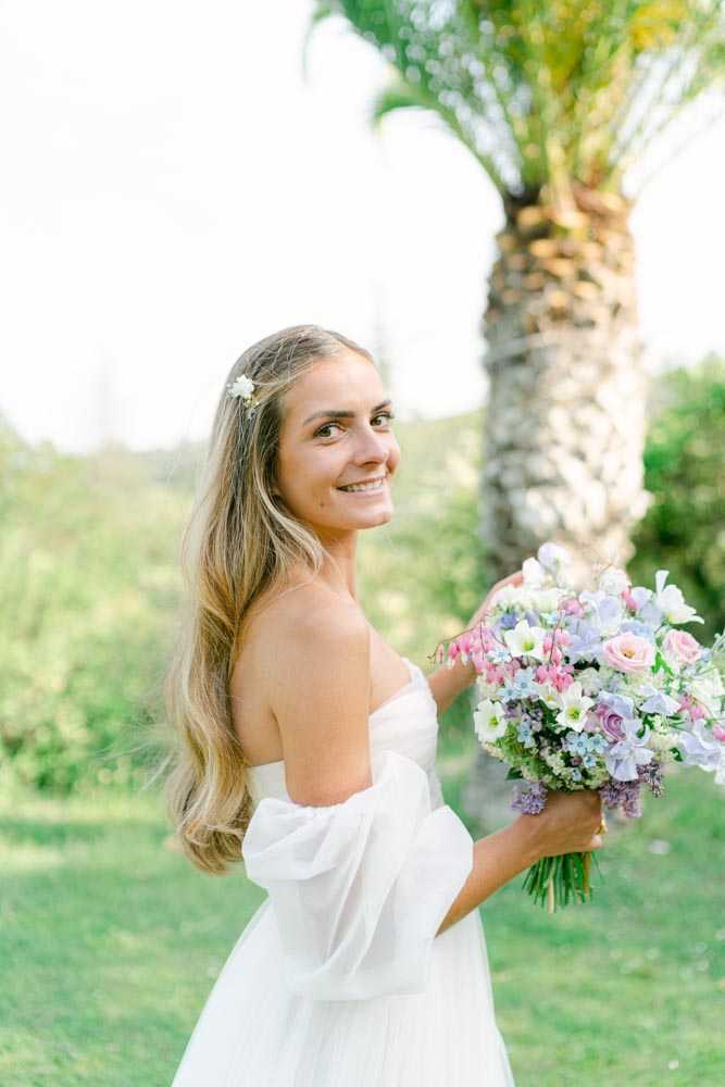 A bridal portrait taken outdoors on a lawn, with the bride glancing back over her shoulder toward the camera. She wears a white off-the-shoulder dress with voluminous puff sleeves and has long, loosely worn blonde hair accessorized with a small white floral hair pin. She holds a large, loosely arranged bouquet featuring soft pink sweet peas, mauve roses, pale blue delphinium, white lisianthus, and lavender blooms. The styling is relaxed and romantic with a pastel floral palette. The portrait is shot at a medium distance with a shallow depth of field, softly blurring the palm tree and greenery in the background.