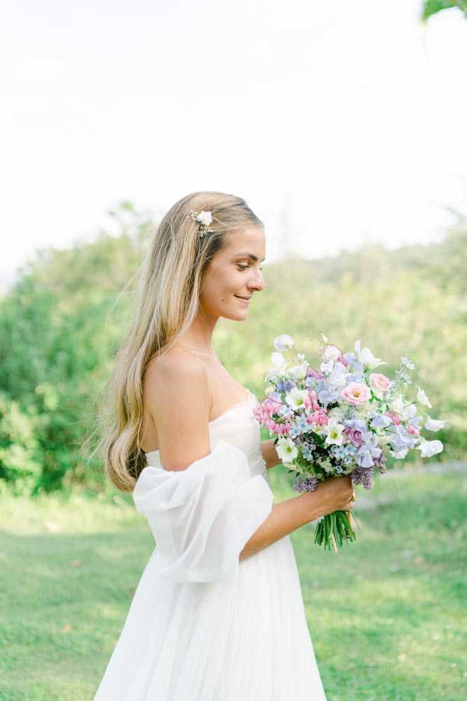 A bridal portrait taken outdoors in a garden setting, showing the bride in a three-quarter profile view looking downward with a slight smile. She wears a white strapless gown with a sheer, billowy off-the-shoulder wrap or sleeves, and her long blonde hair is worn down with a small floral hair pin. She holds a loose, garden-style bouquet composed of pink roses, lavender sweet peas, dusty blue hydrangea, white lisianthus, and small filler blooms with visible green stems. The overall floral palette is soft pastel — pink, lilac, white, and dusty blue. The styling is romantic and light, with a fine-art, airy feel. Medium portrait shot with a shallow depth of field, background softly blurred.