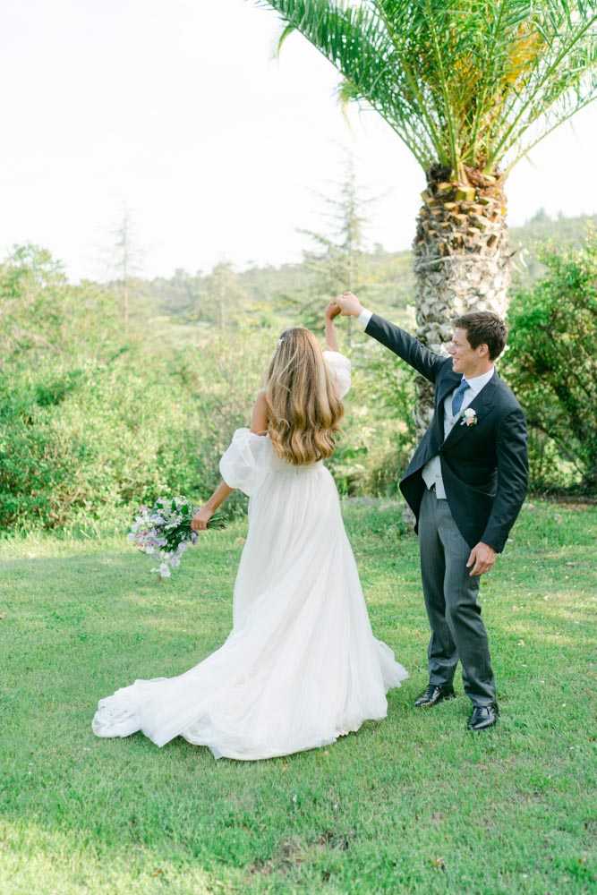 A couple portrait shot outdoors on a lawn, with the groom spinning the bride as she turns away from the camera. The bride wears a white off-the-shoulder gown with voluminous puff sleeves and a flowing train, and carries a loose bouquet of soft lavender and blush blooms. The groom is dressed in a black tailcoat jacket with grey trousers, a light blue tie, and a small white boutonniere. The setting is an open grassy area with a palm tree in the foreground and dense green woodland in the background, suggesting a garden or estate grounds. The shot is a full-length portrait with a bright, airy, light-filled quality consistent with a classic romantic styling.