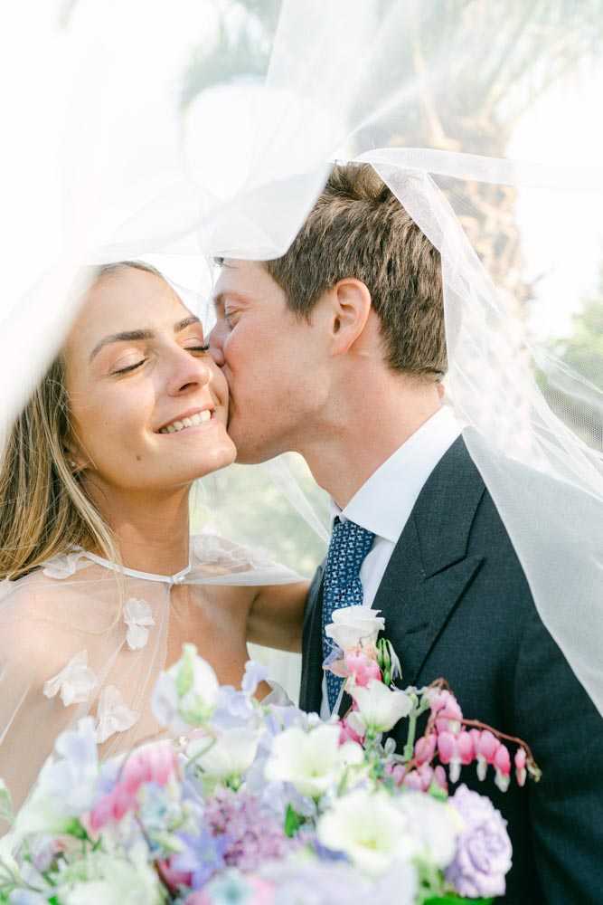 A close-up couples portrait of a bride and groom under the bride's veil, which billows around them and catches bright light. The groom, wearing a charcoal suit with a blue patterned tie and a small floral buttonhole featuring pale pink and white blooms, kisses the bride on the cheek as she smiles with her eyes closed. The bride wears a sheer, halter-style gown with three-dimensional floral appliqués and holds a loose, garden-style bouquet featuring sweet peas in lavender and white, pale pink bleeding heart flowers, white lisianthus, and soft purple clusters. The shot is an intimate, softly lit portrait with a shallow depth of field and an outdoor background visible through the veil.