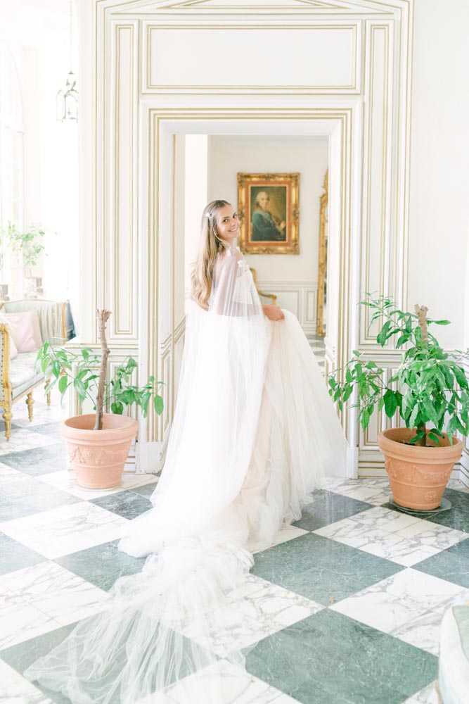 A bride stands alone in the grand interior of what appears to be a French chateau, turning to look back at the camera with a smile. She wears a voluminous ivory tulle ballgown with a sheer tulle cape or overlay, and her long hair is worn down. The room features white-and-gold boiserie wall paneling, a black-and-white marble checkerboard floor, terracotta-potted green plants flanking a doorway, and a gilt-framed oil portrait painting visible through the doorway beyond. A French-style upholstered settee with a blush cushion is partially visible to the left. The overall decor style is classic French period interior. Full-length portrait shot with the dress train spread across the floor. Potential venue feature image.