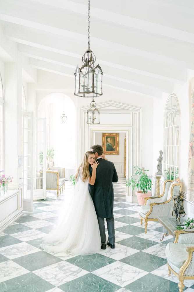 A couple shares an intimate embrace in the grand interior gallery of a French chateau, shot as a full-length portrait from behind. The bride wears a white tulle ball gown with a long flowing train and her hair down, while the groom is dressed in a dark navy suit. The room features a black and white marble checkerboard floor, white paneled walls with gold molding, ornate iron lantern pendant lights, and gilt-framed Louis XVI-style armchairs upholstered in pale blue. A classical oil portrait in a gold frame is visible through a doorway in the background, alongside a stone sculpture and potted green plants. The overall decor palette is white, gold, and soft grey with a classic French interior style. Potential venue feature image.