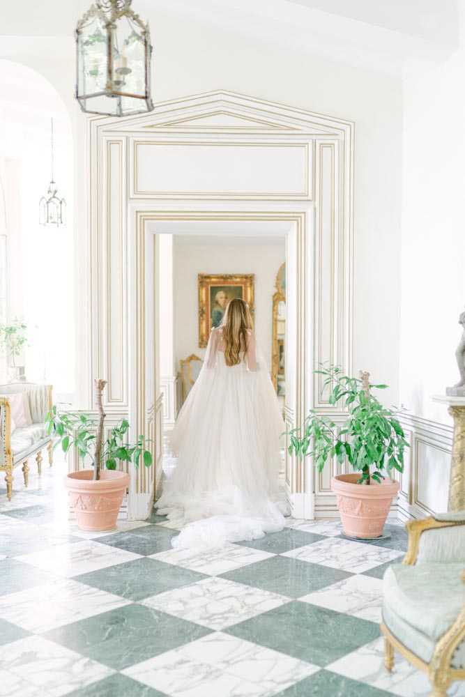 A bridal portrait taken indoors inside a French chateau, showing the bride from behind as she stands in a large doorway with white-painted boiserie paneling trimmed in gold. She wears a voluminous ivory and blush tulle ball gown with a flowing cape or veil trailing behind her. The interior features a black-and-white checkered marble floor, antique gilt-framed artwork and mirrors visible through the doorway, terracotta pots with green potted plants flanking the entrance, Louis XVI-style gilt furniture with pale blue-green upholstery, and iron-and-glass lantern pendants overhead. The overall decor style is classic French heritage with a formal, ornate aesthetic. Wide portrait shot. Potential venue feature image.