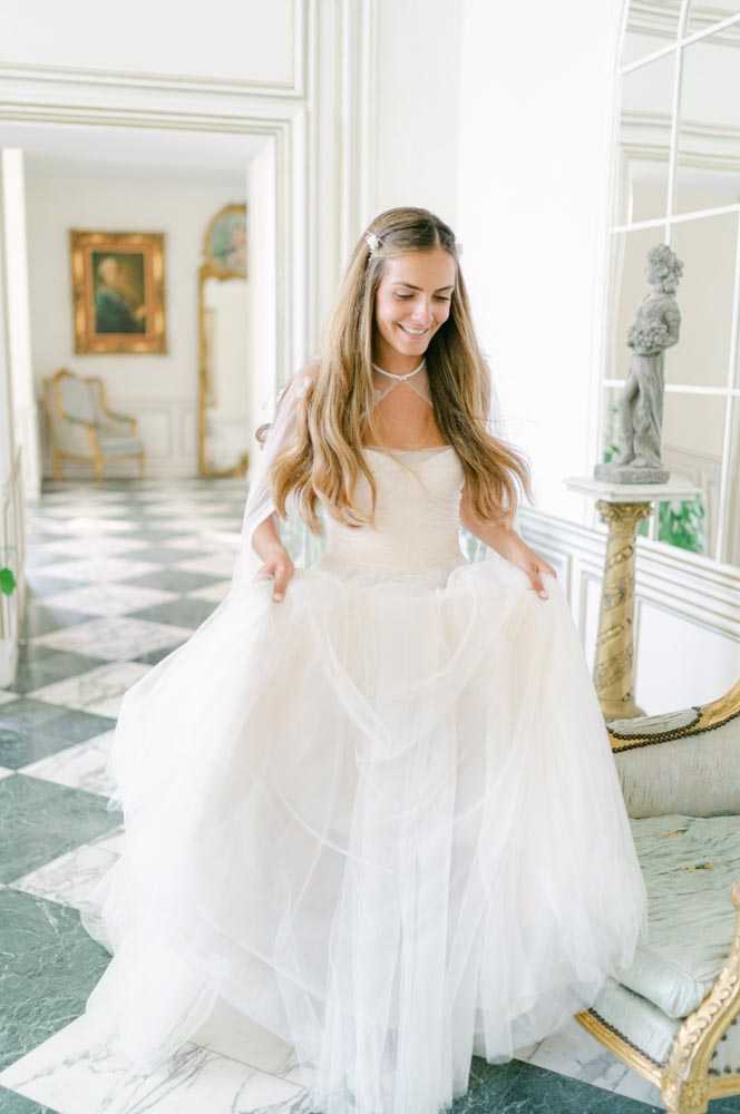 A bridal portrait of a smiling bride holding out the skirt of her white layered tulle ball gown with a sweetheart neckline, photographed indoors in a grand classical French interior. She has long loose brown hair with a small crystal hair accessory and wears a delicate diamond choker necklace. The setting features black-and-white checkered marble floors, white boiserie paneling, gilt-framed oil paintings, Louis XVI-style gilded furniture, and a stone sculptural figure on a pedestal, consistent with a chateau interior. The image is a medium portrait shot with soft, bright natural light.