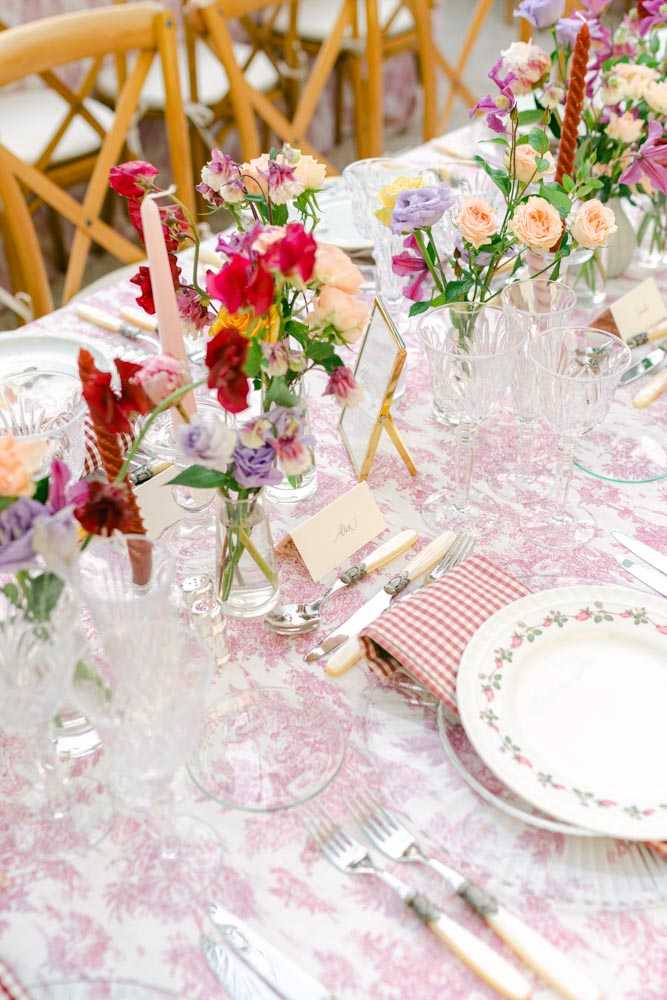 A close-up detail shot of a wedding reception tablescape featuring a pink toile de Jouy tablecloth as the base. The place settings include floral-rimmed china plates with a small rose motif, red-and-white gingham napkins, silver flatware with cream-colored handles, and handwritten calligraphy place cards alongside a small gold easel table number holder. Multiple clear glass bud vases are clustered as centerpieces holding a colorful wildflower-style arrangement of deep red sweet peas, peach garden roses, lavender lisianthus, hot pink blooms, and mixed greenery, accompanied by individual taper candles in blush pink and terracotta red. The overall decor palette is playful and garden-inspired, mixing bold saturated florals with soft pink printed linens and natural wood cross-back chairs visible in the background.