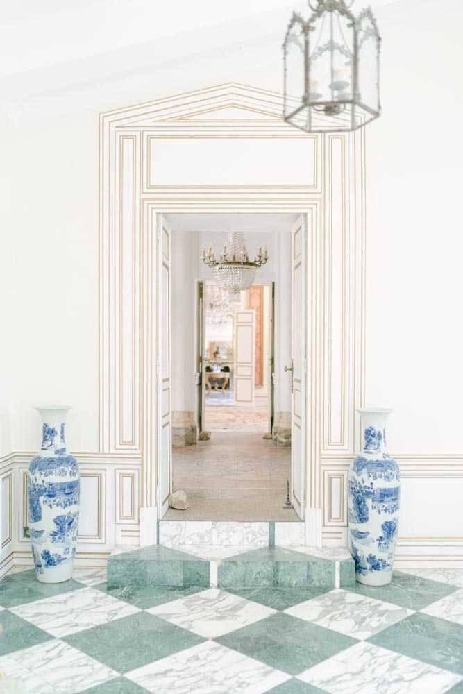 An interior architectural detail shot of a grand venue entrance hall featuring a green and white marble checkerboard floor, white double doors framed by ornate cream and gold paneling with a classical pediment surround. Two large blue and white Chinese-style porcelain floor vases flank the doorway symmetrically. In the foreground, a hexagonal brass and glass lantern hangs from the ceiling, while through the open doors a crystal chandelier is visible in the adjoining room. No people are present. The wide shot captures the formal, classical French interior design style with its gilded wall moldings and enfilade room layout. Potential venue feature image.