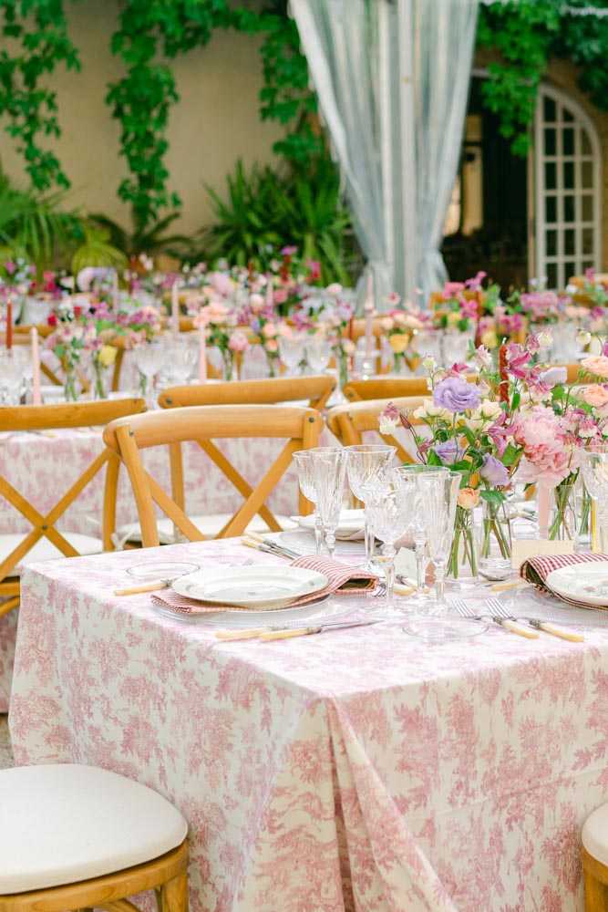 An outdoor wedding reception tablescape detail shot showing multiple long guest tables set in what appears to be a courtyard or garden terrace of a French-style venue with arched windows and ivy-covered walls visible in the background. The tables are dressed in pink toile de Jouy print tablecloths with red-and-white striped napkins, china plates with decorative borders, wood-handled flatware, and cut crystal glassware. Centerpieces consist of loose arrangements of blush peonies, lavender lisianthus, pink sweet peas, peach ranunculus, and garden roses displayed in clear glass bud vases of varying heights. Natural wood cross-back chairs are placed at each setting, and soft pink taper candles are visible along the tables in the background. Sheer light blue draping is used as a backdrop element between the tables and the venue facade. The overall decor palette is pink, lavender, peach, and white with a romantic French country styling theme. Wide-angle detail shot taken from a low angle to capture the table depth and room layout.