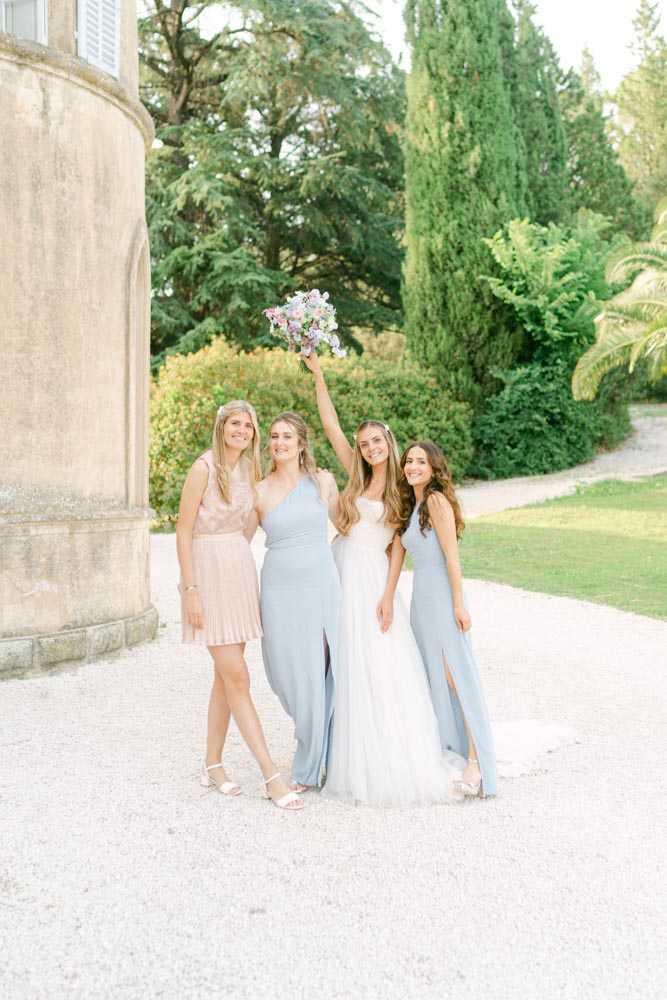A bridal party portrait taken outdoors on a gravel path alongside a chateau. The bride, wearing a white tulle gown with a lace bodice, stands at center holding a bouquet of soft pink and lilac blooms raised above her head, while posing with three bridesmaids. Two bridesmaids wear dusty blue floor-length dresses with a slit — one in a one-shoulder style — and the third wears a short blush pink sequined and pleated mini dress. The group of four women are smiling and posed in a relaxed, celebratory stance. The styling palette is soft and pastel, consistent with a classic French chateau wedding aesthetic. This is a full-length portrait shot in natural light.