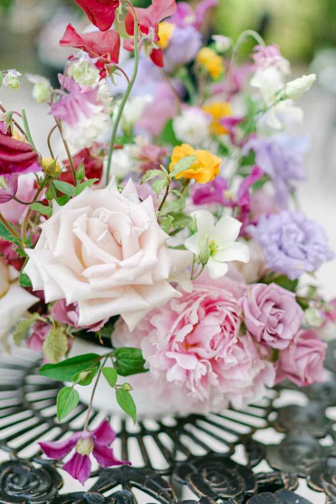 Close-up detail shot of a floral arrangement resting on a dark wrought-iron garden table. The arrangement features a mixed palette of blush pink roses, mauve garden roses, lavender lisianthus, deep magenta sweet peas, orange ranunculus, white star-shaped flowers, and trailing green vine foliage. The style is loose and garden-inspired with an abundant, unstructured form. The background is softly blurred, suggesting an outdoor setting. The overall color palette is vivid and multicolored, consistent with a bohemian or garden-party wedding aesthetic.