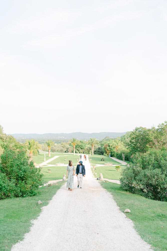 A couple walks hand-in-hand along a wide gravel allée bordered by manicured lawns, palm trees, and lush greenery, with rolling hills visible in the background. The woman wears a long floral dress in white and green tones, and the man is dressed in a navy blazer with light-colored trousers and a hat. Several other guests are visible further down the path in the background. The wide shot is taken from an elevated perspective looking down the allée, giving a strong sense of depth and the formal garden layout of the estate. Potential venue feature image.