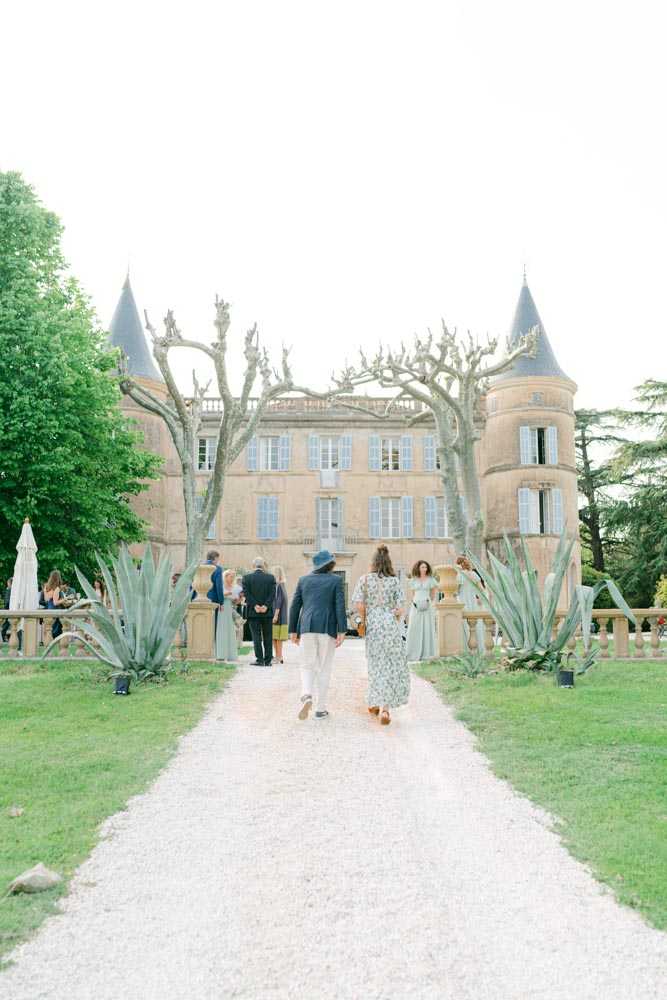 Guests walk along a gravel pathway toward a French chateau during what appears to be a cocktail hour or arrival. The chateau is a three-story stone building with pale blue shutters, two conical-roofed turrets, and a stone balustrade terrace, flanked by large agave plants. In the foreground, two guests walk away from the camera — one in a dark blazer with a blue hat and white trousers, the other in a floral midi dress — while approximately eight additional guests in smart-casual attire gather near the terrace. Wide shot taken from ground level along the central pathway axis. Potential venue feature image.