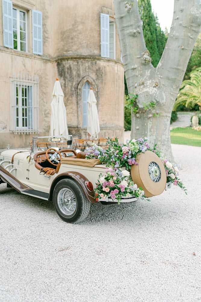 A cream and brown vintage convertible wedding car is parked on a gravel driveway in front of a French chateau with light blue shutters and a distinctive rounded tower. The rear of the car is decorated with an abundant floral arrangement featuring mauve, blush pink, white, and lavender blooms — likely roses and delphinium — with lush green foliage cascading around the spare tire mounted on the back. The car interior features tan leather upholstery and a wooden steering wheel. No people are visible in the frame; this is a detail/wide shot focused on the decorated vehicle with the chateau facade as backdrop. Potential venue feature image.
