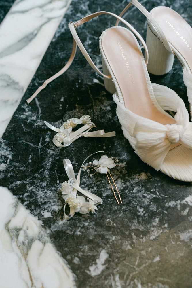 A flat lay detail shot of bridal accessories arranged on a dark veined marble surface. The centerpiece is a pair of Loeffler Randall ivory block-heel sandals with a pleated bow knot detail and ankle strap. Alongside the shoes are two small floral hair accessories — white ribbon-tied clusters featuring ivory fabric or dried flowers with pearl and bead details — and a single gold bobby pin topped with a small white floral and pearl embellishment. The overall accessory palette is ivory, cream, and gold, consistent with a classic or modern bridal styling approach.