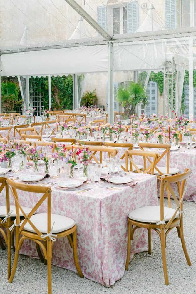 A wedding reception setup photographed in a wide shot inside a clear-roof marquee tent erected in the courtyard of what appears to be a French chateau or manor house, visible through the transparent walls with pale blue shutters and ivy-covered facade. Multiple long rectangular tables are dressed in pink and white toile de Jouy print tablecloths, set with patterned china plates, pink striped napkins, glassware, and gold cutlery. Clusters of bud vases filled with loose arrangements of pink, purple, magenta, and peach flowers — including sweet peas, roses, and ranunculus — run as low centerpieces down each table. Natural wood cross-back chairs with white cushion pads are positioned throughout, and thin taper candles in warm tones are interspersed among the florals, contributing to a romantic, garden-party aesthetic with a French country styling theme.