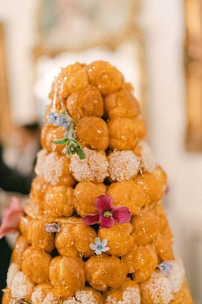 Close-up detail shot of a traditional French wedding croquembouche — a tower of caramel-glazed choux pastry puffs stacked in a cone shape and decorated with small fresh flowers including a deep pink bloom, pale blue forget-me-nots, and a tiny white flower. Alternating tiers feature puffs coated in crushed praline or nougatine, adding textural contrast. Caramel threads are visible draping over the structure. The background is softly blurred but shows a gilded gold mirror frame and pale interior walls consistent with a classic French interior, likely a chateau reception room. The shot is tightly cropped on the dessert with shallow depth of field.