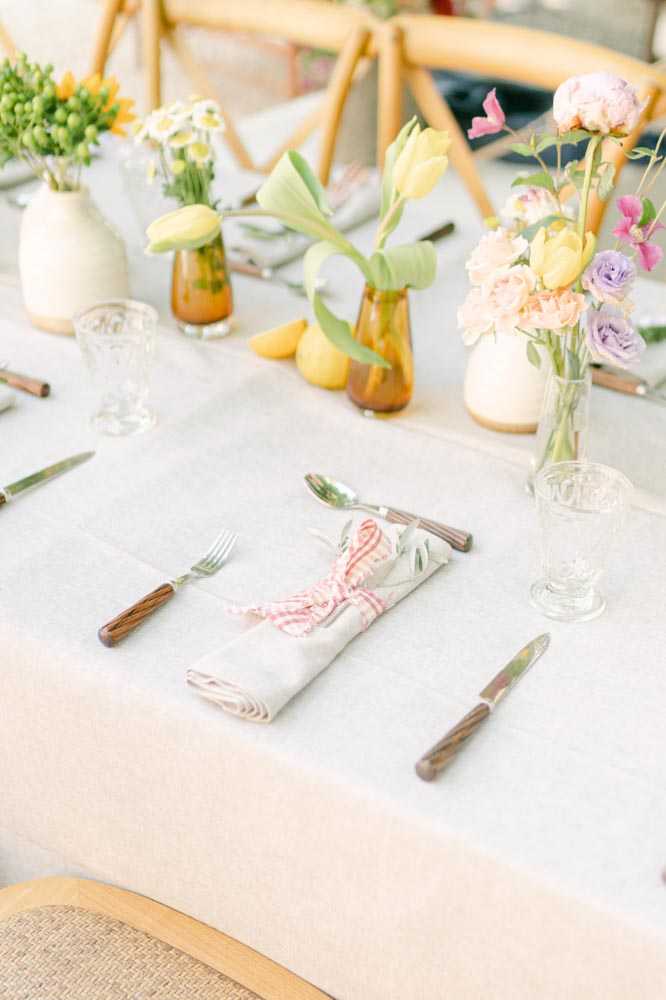 Close-up detail shot of a wedding reception table setting with a soft white linen tablecloth. The place setting features a folded linen napkin tied with a pink gingham ribbon, flanked by wood-handled cutlery in a fork-knife arrangement, alongside clear pressed-glass tumblers. The centerpiece styling uses a mix of small cream ceramic vases and amber glass bud vases holding yellow tulips, white daisies, and green button flowers, alongside a larger clear glass vase with a loose arrangement of peach garden roses, lavender roses, yellow tulips, and pink sweet peas; fresh lemons are scattered between the vases as decorative accents. The overall palette is light and springlike — pastel multicolor florals against neutral linens — with natural wood cross-back chairs visible in the background, suggesting a relaxed, garden-party aesthetic.