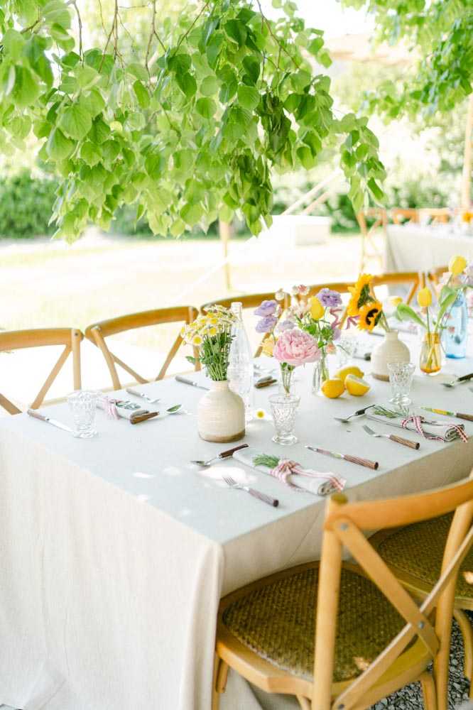 A close-up detail shot of an outdoor wedding reception table set under a leafy tree canopy, likely within a tent or open-air structure. The long table is dressed with a white linen tablecloth and set with wooden-handled cutlery bundled in white napkins tied with small red-and-white gingham ribbon. Place settings include crystal glassware and smaller cut-crystal glasses. The centerpiece decor features an assortment of small bud vases and ceramic vessels holding mixed floral arrangements including a pink peony, lavender blooms, white daisies, yellow tulips, and a sunflower, alongside loose lemons scattered along the table surface. Natural wood cross-back chairs surround the table. The overall styling is relaxed and garden-inspired with a cheerful, colorful palette of yellow, pink, lavender, and white against neutral linens.