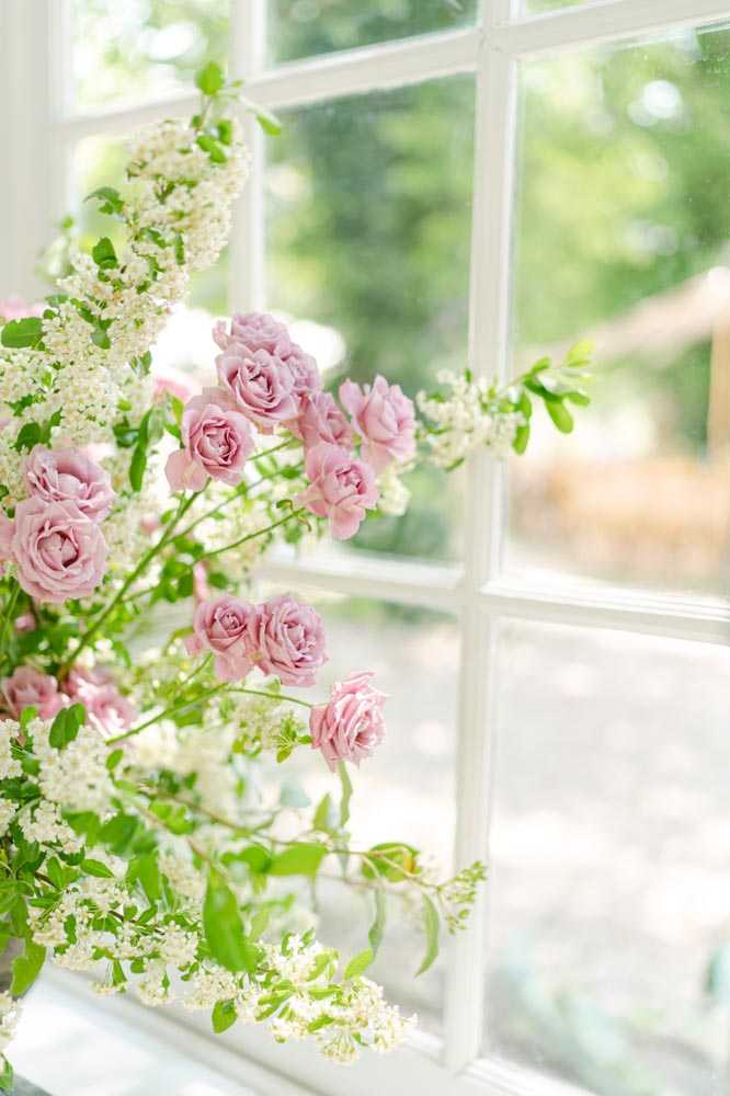 Close-up detail shot of a floral arrangement placed on or near a white-framed window ledge, with soft natural light coming through the glass. The arrangement features soft pink garden roses and clusters of small white flowers, likely spirea or similar delicate filler blooms, with fresh green foliage throughout. The loose, garden-style arrangement has an unstructured, organic form with stems extending outward in multiple directions. The background is softly blurred, suggesting an indoor setting with a view of greenery outside.