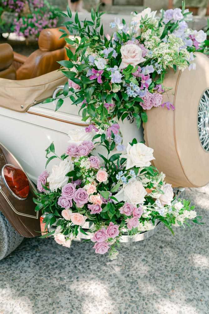 Detail shot of the rear of a vintage cream-colored convertible wedding car decorated with an abundant floral arrangement. The flowers include mauve and lavender roses, blush pink roses, ivory roses, light blue delicate blooms, small white filler flowers, and lush green foliage that cascades down from the trunk and spare tire mount. The arrangement is full and trailing, with stems and leaves draping toward the ground. The car features tan leather interior visible in the background and chrome detailing. The floral palette is a soft mix of purple, mauve, blush, and white, consistent with a romantic garden-style wedding aesthetic.