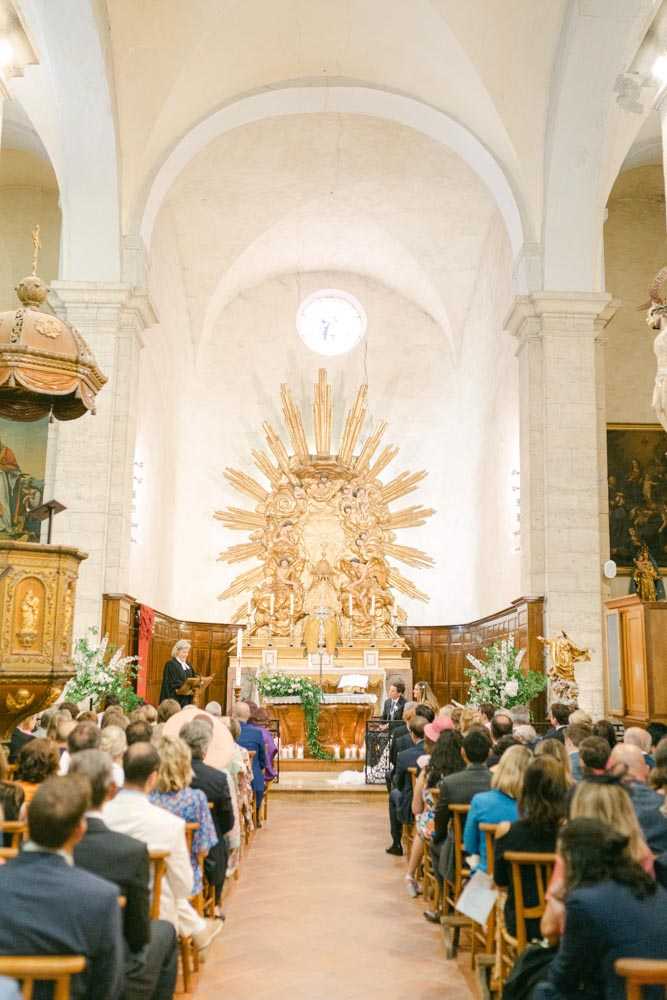 A religious wedding ceremony taking place inside a French stone chapel with white vaulted ceilings and barrel arches. The altar is dominated by a large ornate gilded sunburst retable with baroque sculptural detail, flanked by floral arrangements of white blooms and greenery on each side. Approximately 80–100 guests are seated in wooden chairs arranged in two sections facing the altar, dressed in smart-casual to formal attire in a mix of navy, white, and colorful summer dresses. The shot is a wide, central-aisle perspective from the rear of the chapel, capturing the full interior architecture and the ceremony in progress at the altar.