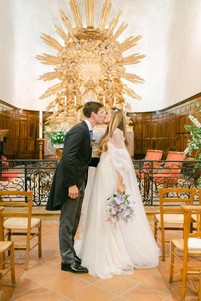 The bride and groom share their first kiss at the altar inside a small historic chapel with terracotta tile floors, dark wood paneling, and a large ornate gilded sunburst altarpiece as the backdrop. The groom wears a charcoal grey suit with a blue tie, while the bride wears a white tulle gown with a sheer cape overlay and a floral hair accessory. The bride holds a loose, garden-style bouquet in shades of lavender, soft pink, and white. Simple wooden rush-seat chairs line the aisle, and white floral arrangements are visible near the altar; the overall styling is romantic and classic. This is a mid-range portrait shot capturing the couple full-length in the center of the chapel.