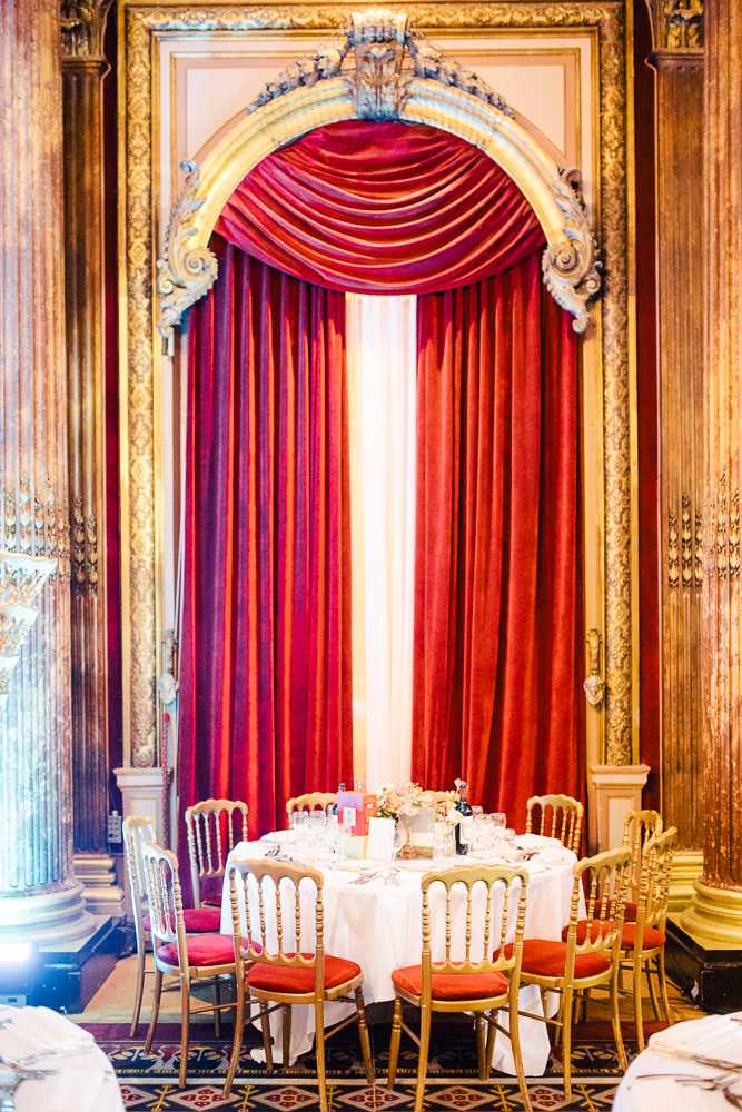 A wedding reception table setup photographed in a formal indoor ballroom with ornate gilded architectural details, including carved pilasters and an elaborate arched molding framing floor-to-ceiling deep red velvet drapes. A single round dining table is set with a white linen tablecloth, glassware, place settings, and a small low floral centerpiece featuring light pink blooms. The chairs are gold Chiavari-style with red cushioned seats, arranged around the table for approximately eight guests. The room features decorative tile flooring and warm amber-toned wall paneling, creating a classic, formal décor palette of gold, red, and white. The shot is a medium wide portrait-orientation image centered on the table and drapery backdrop. Potential venue feature image.