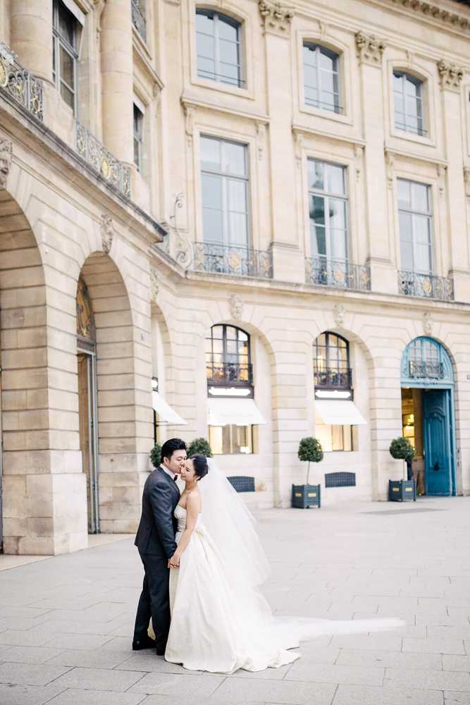 A couple portrait taken outdoors in a Parisian square, identifiable by the classical limestone Haussmann-style architecture with ornate wrought-iron balconies and arched windows in the background. The bride wears a strapless ivory ball gown with a long cathedral-length veil trailing across the cobblestone pavement, while the groom wears a charcoal suit with a dark tie; the two stand close together in an almost-kiss pose. The setting appears to be Place Vendôme or a similar grand Parisian square, with a teal-painted arched doorway and topiary trees visible in the background. The composition is a full-length portrait shot with the couple positioned left of center, emphasizing both the architectural backdrop and the sweep of the bride's gown and veil.