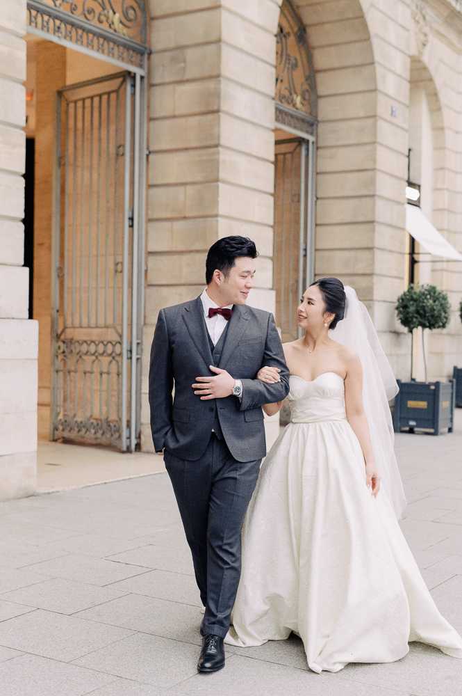 A couple portrait taken outdoors in front of a classical Parisian limestone building with ornate wrought-iron gates and arched doorways. The bride and groom are walking together, with the bride linking her arm through the groom's as they look at each other. The groom wears a charcoal grey three-piece suit with a burgundy bow tie and black dress shoes. The bride wears a strapless ivory ball gown with a structured bodice and a cathedral-length veil, with her hair swept up. The shot is a full-length portrait with the architectural facade as the background.