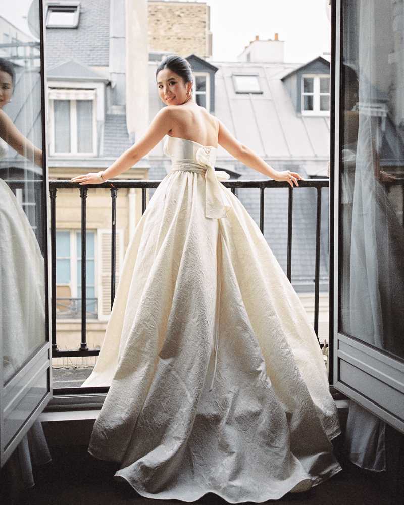 A bride stands at an open French balcony door in what appears to be a Parisian hotel room, with classic Haussmann-style rooftops and zinc mansard roofs visible in the background. She is posed with her back to the camera, looking over her shoulder with a smile, wearing a strapless ivory ball gown with a jacquard or brocade texture and a large satin bow at the back waist. Her dark hair is styled in a sleek updo and she wears a choker necklace. The composition is a full-length portrait shot, framed symmetrically by the two open window panels, with sheer curtains on either side and a partial reflection of the bride visible in the left window glass.
