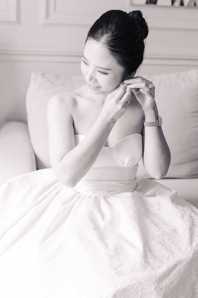 Black-and-white portrait of a bride getting ready, seated on a light-colored sofa or chaise in what appears to be an indoor bridal suite with panelled walls. She is putting on an earring, with both hands raised near her ear, and wears a strapless structured bodice wedding gown with a full skirt visible in the lower frame, along with a metal bracelet or watch on her wrist. Her dark hair is styled in a neat high bun. The image is a close-to-medium portrait shot with soft, bright tones and gentle contrast, creating an airy, light-filled mood.