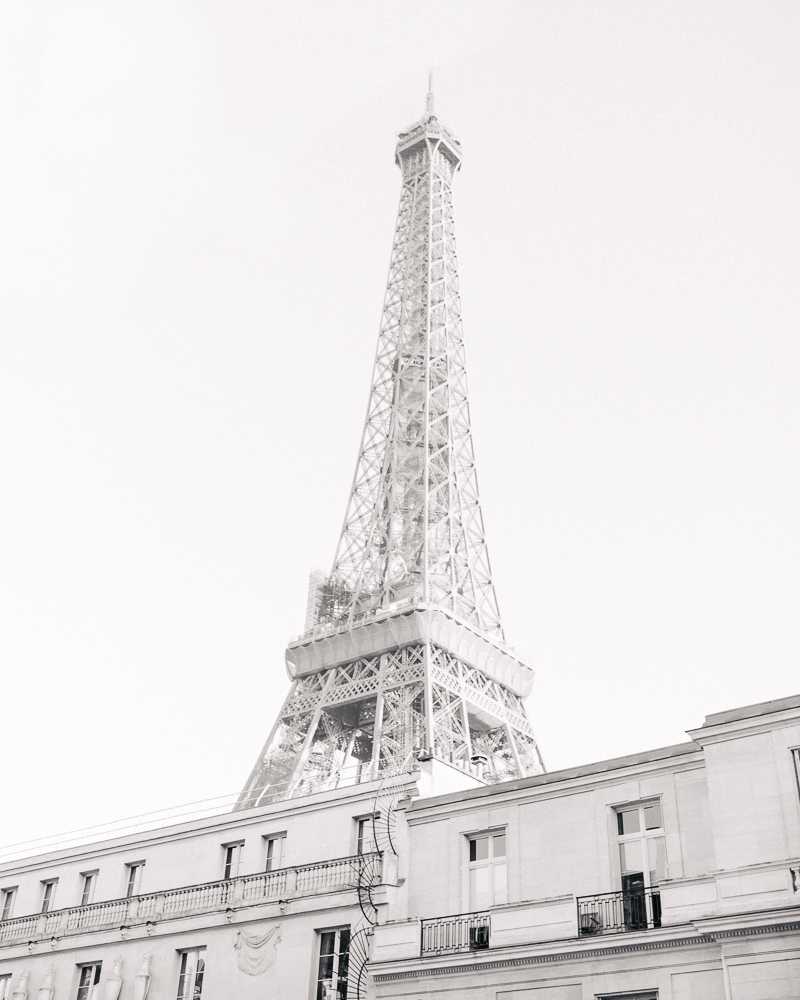 This is a high-contrast black-and-white architectural shot taken in Paris, looking upward from street level past a classic Haussmann-style building with wrought-iron balconies and decorative facade details toward the Eiffel Tower rising prominently above it. No people or wedding party are visible in the frame. The image is a wide-angle, low-angle composition with bright, blown-out highlights giving it an airy, washed-out tonal quality. This appears to be a Paris location establishing shot, likely used as a backdrop context image for a Parisian wedding. Potential venue feature image.