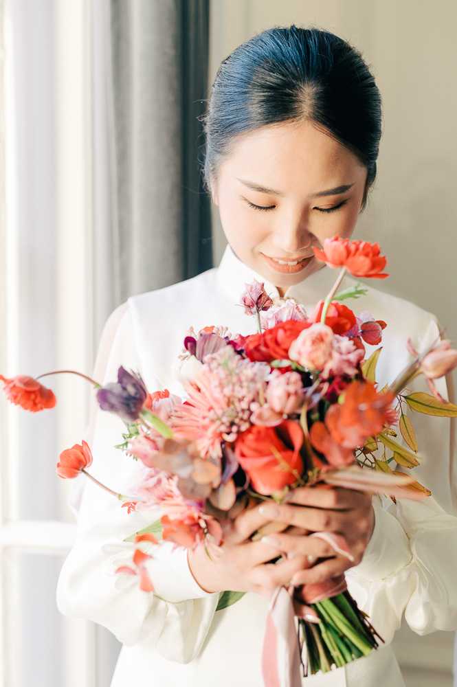 A close-up portrait of a bride holding her bouquet indoors, photographed in front of a softly lit window with neutral curtains in the background. She wears a white high-neck dress with long sleeves, consistent with an ao dai or similar traditional-inspired silhouette, and her dark hair is pulled back in a sleek low bun. The bouquet is a vibrant mix of coral and orange poppies, red roses, blush pink dahlias or chrysanthemums, purple tulips, and assorted wildflower-style stems with foliage, tied with a pale pink ribbon. The bride looks downward toward the flowers with a gentle smile, and the composition is a portrait-style shot with shallow depth of field that keeps focus on her face and the bouquet.