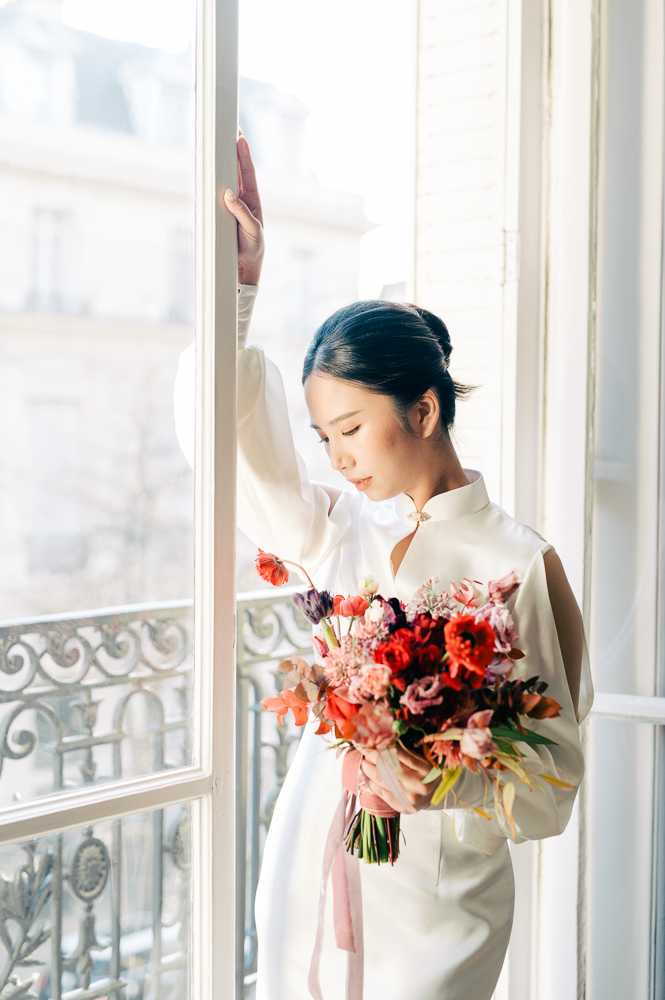 A bridal portrait of a woman standing beside tall French windows with a wrought-iron Haussmann-style balcony visible in the background, suggesting a Parisian apartment setting. She wears a white satin qipao-inspired dress with a mandarin collar, cold-shoulder cutouts, and long sleeves, paired with a gold collar accessory. Her dark hair is pulled back into a low bun, and she looks downward with one hand resting on the window frame. She holds a lush, loosely arranged bouquet featuring red dahlias, orange and red tulips, mauve lisianthus, dusty pink blooms, and trailing pink silk ribbons. The image is a medium portrait shot with bright natural window light creating a soft, airy atmosphere.