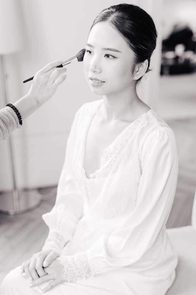 Black-and-white portrait of a bride during the getting-ready stage, seated while a makeup artist applies powder to her cheek with a large brush. The bride wears a white long-sleeved robe with lace trim at the cuffs and neckline, and her dark hair is pulled back in a sleek updo. The image is shot in a bright, softly lit indoor room with a mirror visible in the background. The composition is a close-up portrait with high contrast and bright highlights, emphasizing the bride's calm expression and refined makeup application.