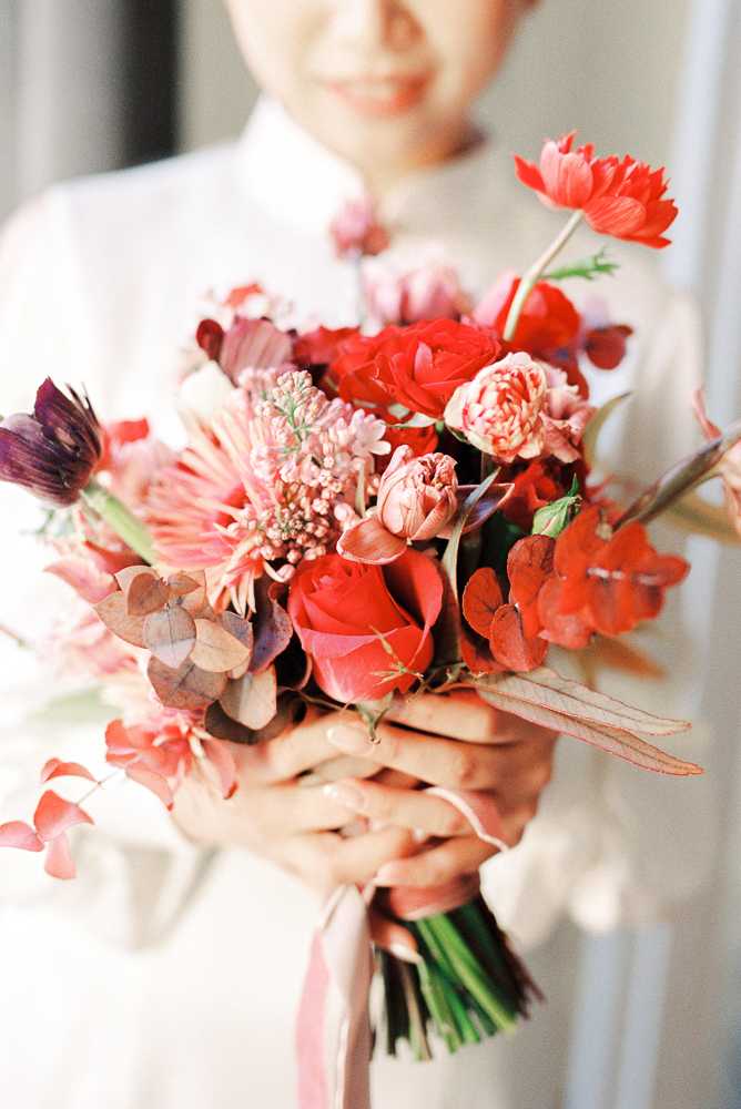 Close-up portrait shot of a bride holding her bouquet toward the camera, with the bride softly out of focus in the background wearing a white high-collar dress. The bouquet is the focal point and features a rich mix of red roses, blush garden roses, red poppies or tulips, mauve dahlias, burgundy foliage, dusty pink waxflower, small blush carnations, and deep red anthurium-style leaves, creating a bold red-to-blush palette with dark burgundy accents. The stems are wrapped and tied with a soft blush pink ribbon. The overall floral style is lush and garden-inspired with a rich, romantic color palette.