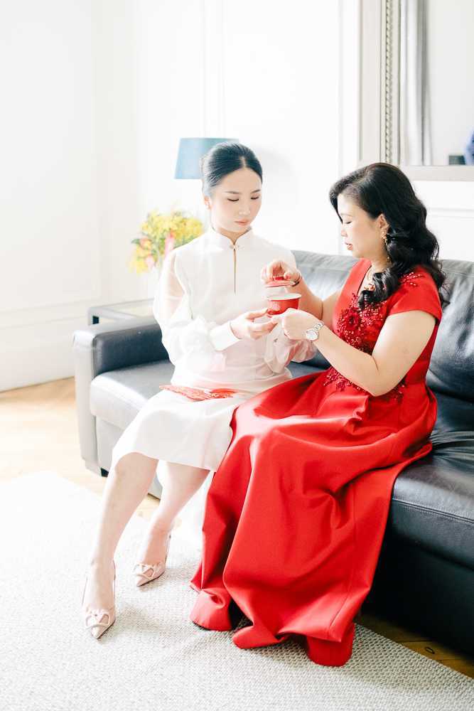 A Chinese tea ceremony moment captured indoors in a bright, white-walled room, showing two women seated together on a dark leather sofa. The bride wears a white qipao-style dress with a mandarin collar and embroidered hem detail, paired with blush pointed-toe heels with bow embellishments, her hair worn in a sleek low bun. The older woman, likely the bride's mother, wears a bright red floor-length dress with cap sleeves and floral beaded embroidery at the neckline. The two are focused on a red lacquered tea cup and lid being exchanged between them, a traditional element of the Chinese wedding tea ceremony. A small arrangement of yellow and coral flowers is visible in the background alongside a navy lamp. The shot is a medium full-length portrait with soft, bright natural light from a nearby window.
