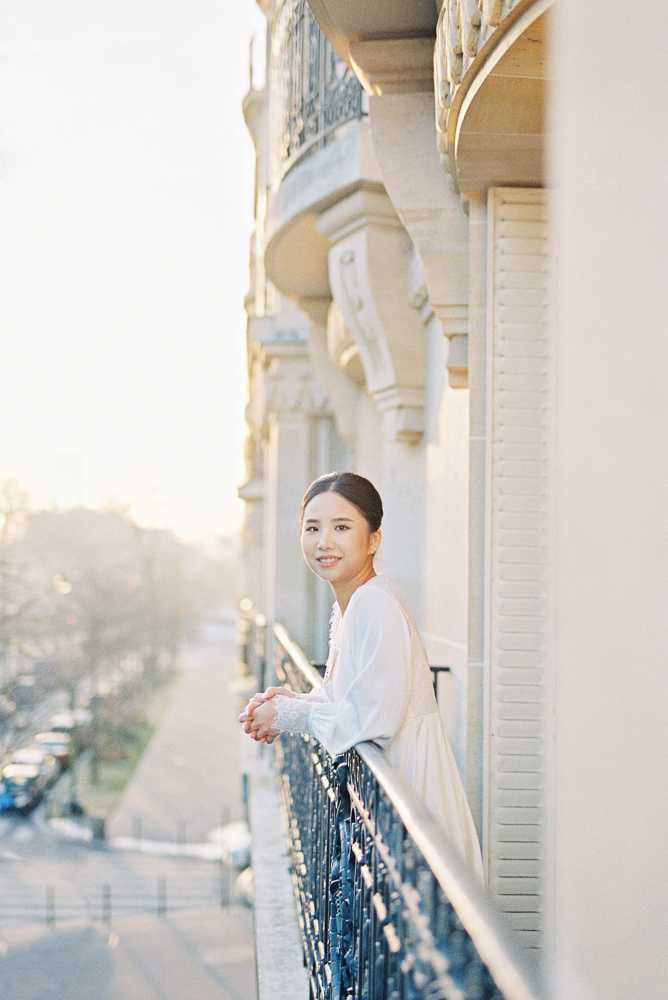 A bride stands on an ornate wrought-iron balcony of a classic Haussmann-style building, leaning on the railing and looking back toward the camera with a relaxed smile. She wears a white long-sleeve bridal top or gown with delicate ruffle or lace cuff detailing, and her dark hair is pulled back in a low updo. The shot is taken during golden hour, bathing the cream stone facade and repeating arched balconies in warm, soft light. This is a portrait composition with a shallow depth of field, with a street and parked cars visible in the soft-focus background below.