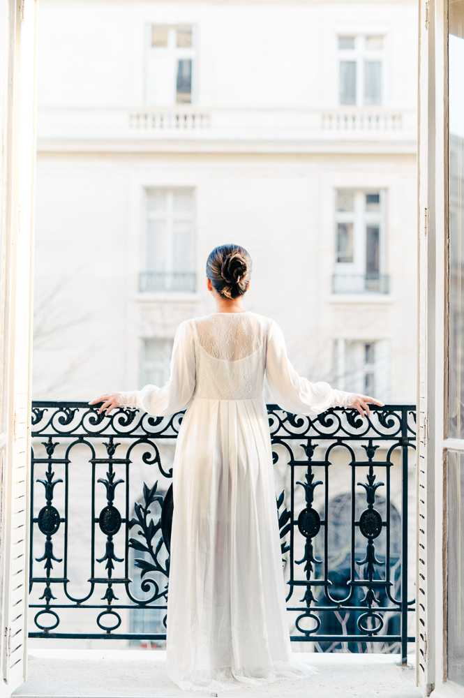 A bride photographed from behind stands on a Haussmann-style apartment balcony in what appears to be Paris, her hands resting on a dark wrought-iron railing with ornate scrollwork and floral motifs. She wears a long-sleeved ivory wedding dress with a sheer lace back panel and flowing chiffon skirt, her dark hair styled in a low bun. The shot is framed by open white French doors, with the cream façade of a classic Parisian building visible and softly out of focus in the background. The image is a full-length portrait with bright, airy natural light and a clean, classic bridal aesthetic.