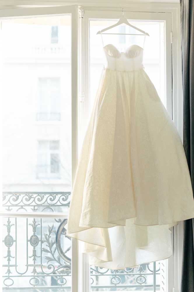 A detail shot of a strapless ivory ball gown wedding dress hanging from a white hanger in front of a large window in what appears to be a Parisian apartment or hotel room. The dress features a sweetheart neckline with structured bodice and a full, voluminous skirt with a subtle textured or jacquard fabric and a tulle underlayer. The window behind the dress shows a classic wrought-iron Haussmann-style balcony railing and the facade of a neighboring building, placing the setting clearly in Paris. The image is backlit by bright natural light, creating a soft, high-key look with a grey curtain visible on the right edge of the frame.