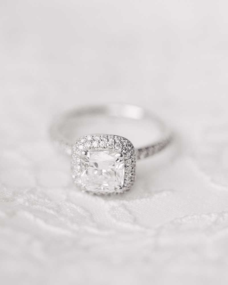 Close-up detail shot of an engagement ring resting on white lace fabric, likely a wedding dress or veil. The ring features a large cushion-cut center stone set in a white gold or platinum halo setting with pavé-set round diamonds surrounding the center stone and continuing down the band on both sides. The squared halo design gives the ring a classic, structured silhouette. The soft, bright lighting and shallow depth of field keep focus sharply on the ring while the lace background provides delicate texture in the foreground and the backdrop fades to a pale grey.