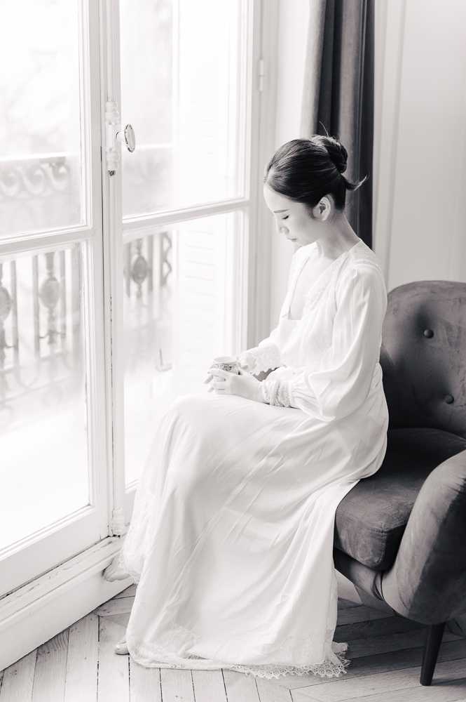 This is a black-and-white getting-ready portrait of a bride seated on a tufted armchair beside large French doors with an ornate wrought-iron balcony visible beyond the glass. The bride wears a long-sleeved, floor-length white robe or bridal cover-up with lace trim at the hem and cuffs, and her dark hair is styled in a neat updo. She is looking downward at a small object held in her hands, suggesting a quiet, reflective moment before the ceremony. The image is a soft full-length portrait with high-key lighting, light tones, and gentle contrast, shot indoors in what appears to be a Parisian apartment or hotel room.