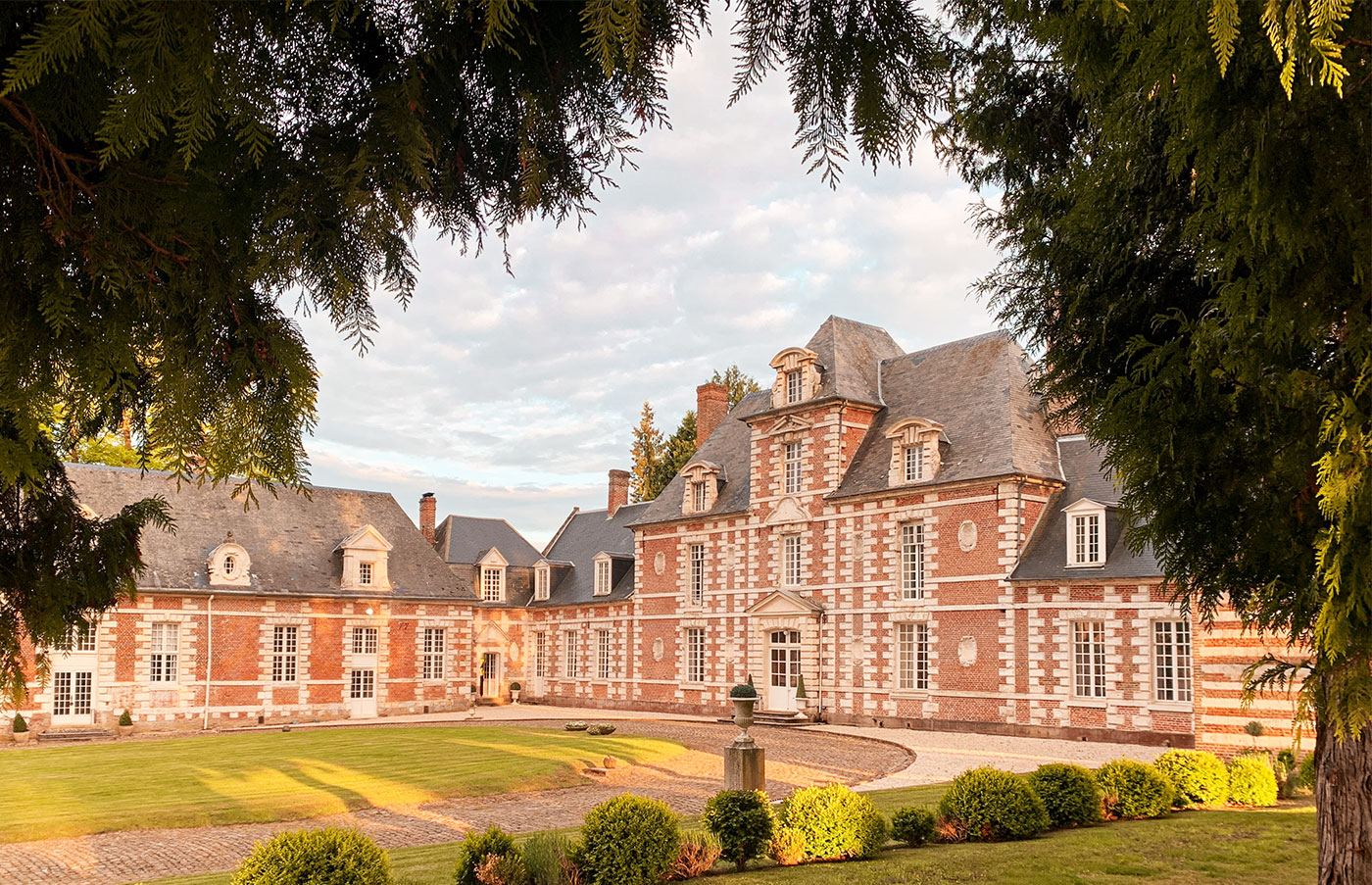 Wide exterior shot of a French château venue photographed at golden hour, with no people present. The building features classic 17th–18th century Norman architecture with red brick and white stone quoining, slate mansard roofs, dormer windows, and multiple chimneys across a multi-wing façade. The forecourt includes a circular gravel driveway with a central stone urn pedestal, clipped boxwood topiaries, and a manicured lawn bathed in warm late-afternoon light. The composition is framed by large conifer tree branches in the foreground, giving the shot depth. Potential venue feature image.