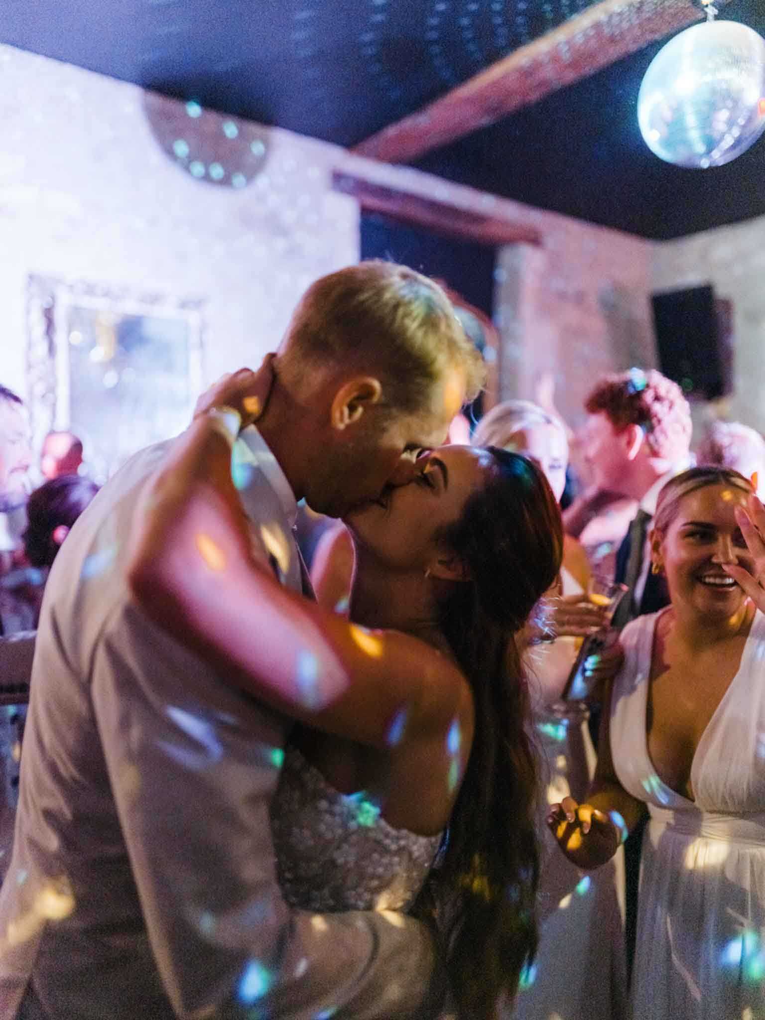 The couple shares a kiss on the dance floor during the evening reception in what appears to be a rustic indoor venue with exposed stone walls and dark-painted ceiling beams. A mirrored disco ball overhead casts scattered light reflections across the scene, contributing to the party atmosphere. The bride wears a fitted embellished strapless gown and the groom is in a light-colored suit, while a guest nearby in a white deep-V dress holds a champagne glass and smiles. The shot is a close-up portrait taken at mid-distance with soft bokeh on the surrounding crowd of guests visible in the background.